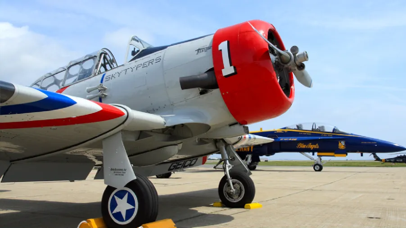 Close-up of a polished radial-engine aircraft with a red nose in the foreground and U.S. Navy Blue Angels jet in the background on the runway