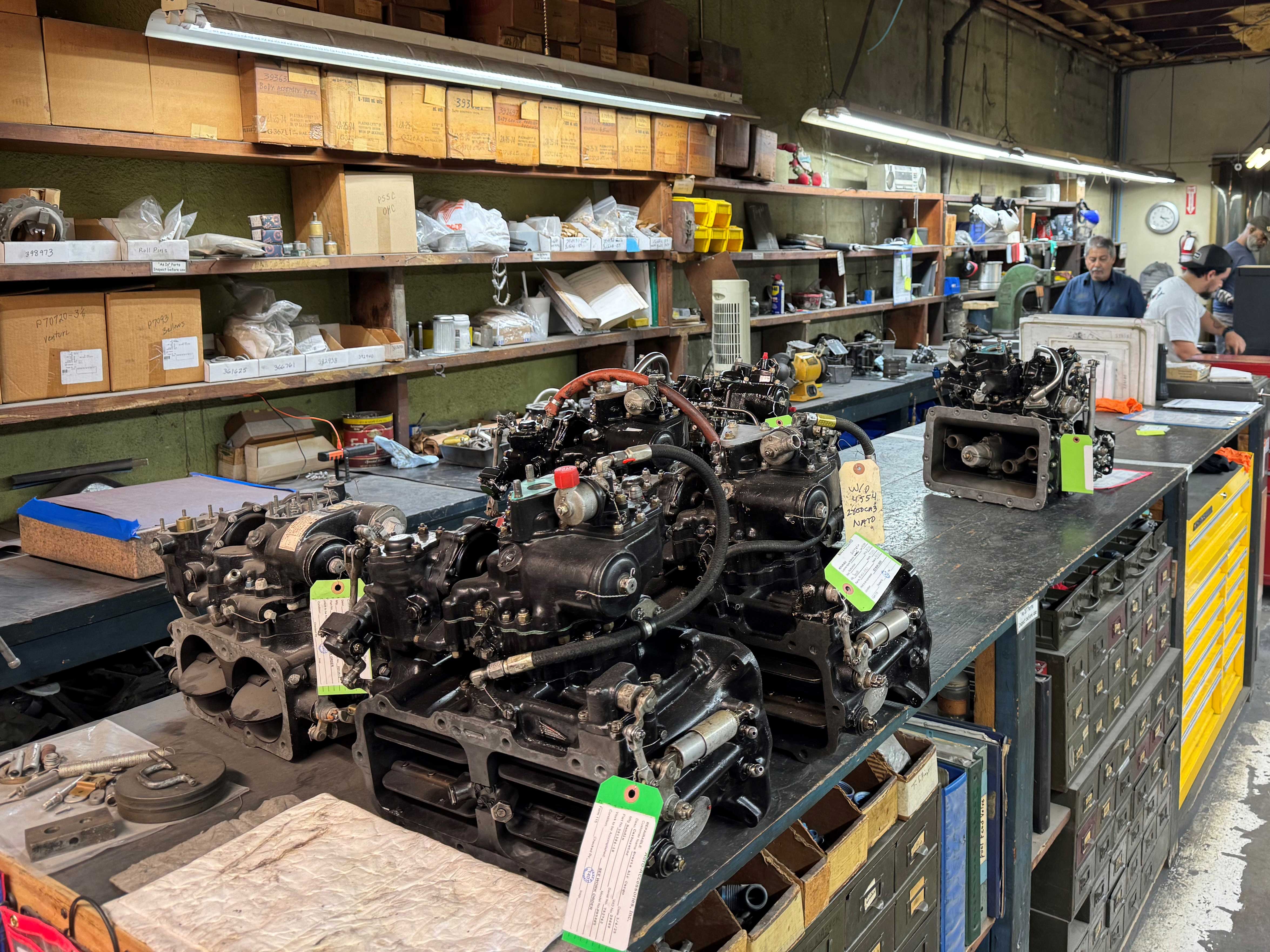 Disassembled aircraft engine components with green inspection tags on a workbench in an MRO shop with parts shelving and technicians in the background