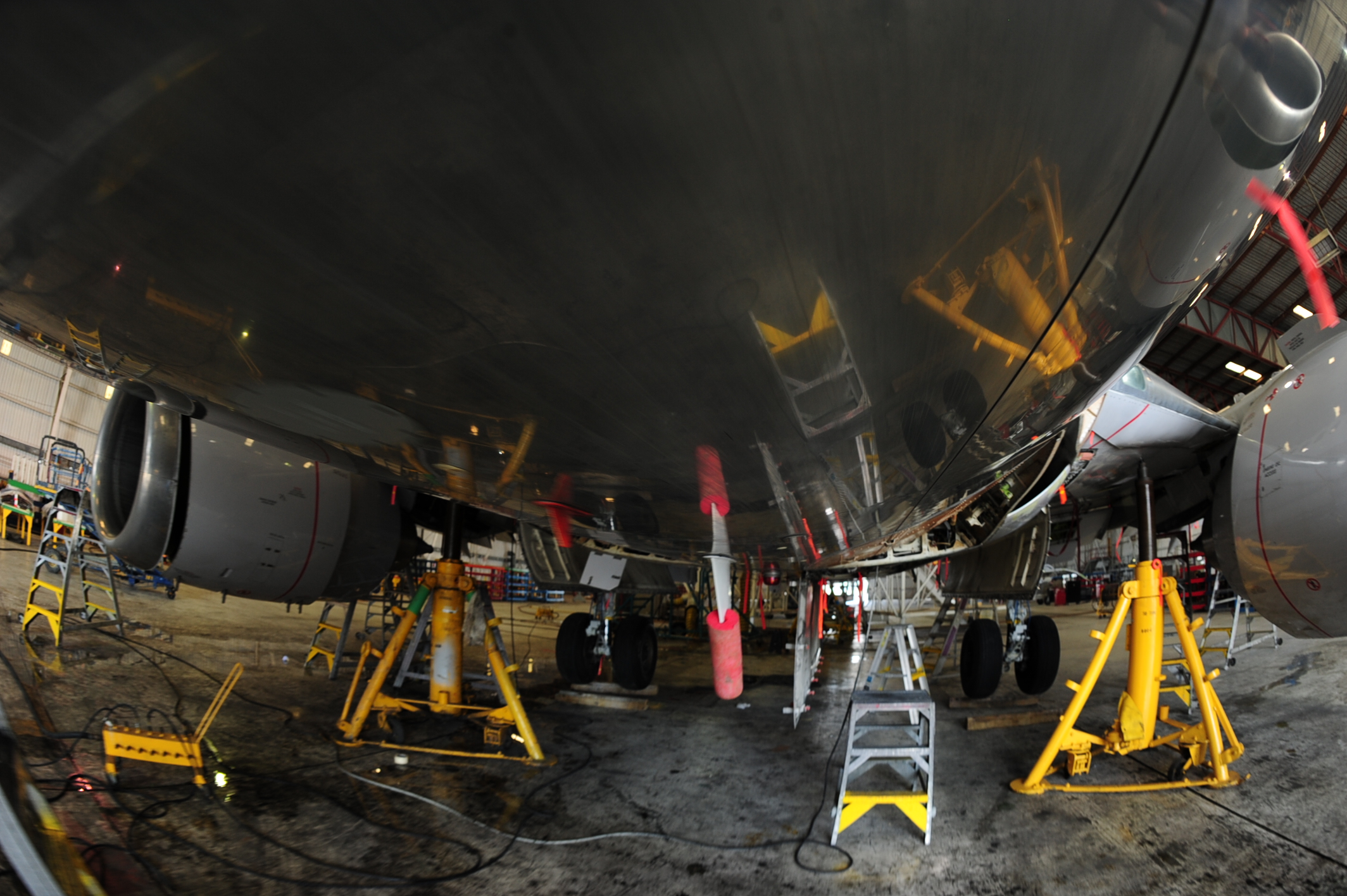 Underside of a commercial airliner raised on yellow maintenance jacks inside an MRO hangar, surrounded by ladders and service equipment