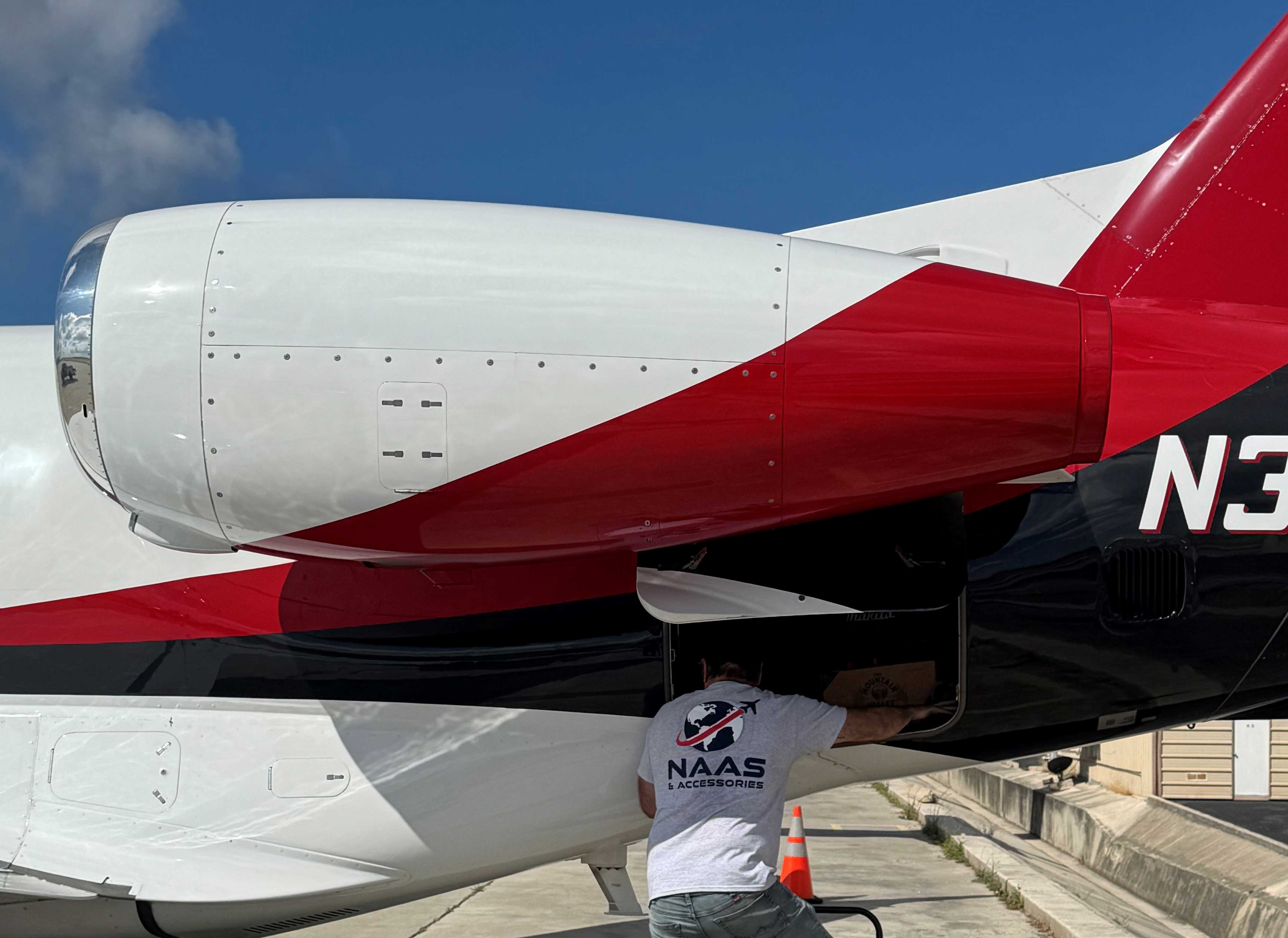 NAAS & Accessories technician inspecting the aft baggage compartment of a red, white, and black corporate jet on the tarmac