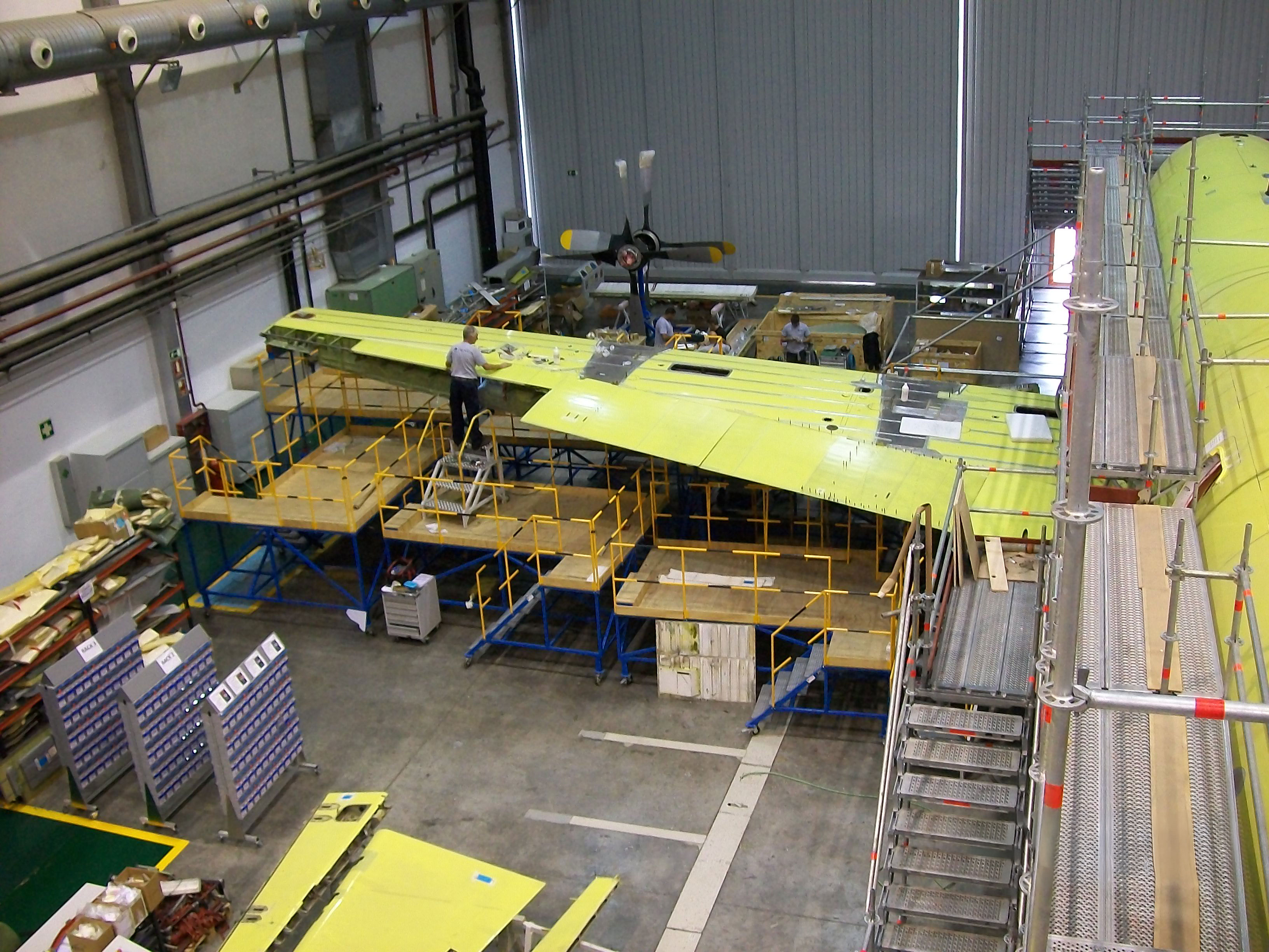 A NAAS & Accessories MRO technician on yellow scaffolding inspecting a large green primer-coated aircraft wing section in an MRO hangar with tools and parts nearby