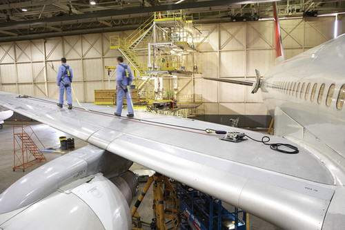 Two maintenance technicians in blue coveralls walking on an aircraft wing inside an MRO hangar with scaffolding and jet engine visible